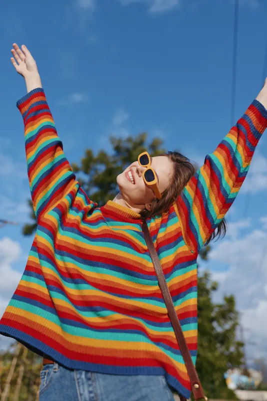 Young woman in her 20s wearing a rainbow sweater and yellow sunglasses, arms raised smiling outdoors under blue sky, casual outfit with curly hair, joyful lifestyle and fashion vibe.