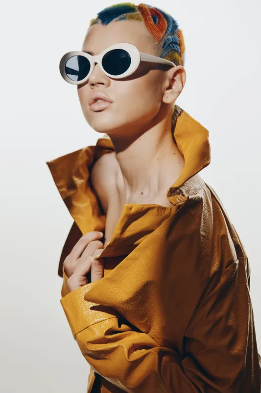 Stylish woman with colorful short hair and white sunglasses wears a mustard yellow jacket, striking a bold and modern pose with a minimal light background.