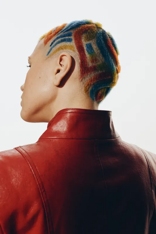 Woman with colorful short hair design and shaved patterns, wearing red leather jacket, seen from behind against white background, creative modern style.