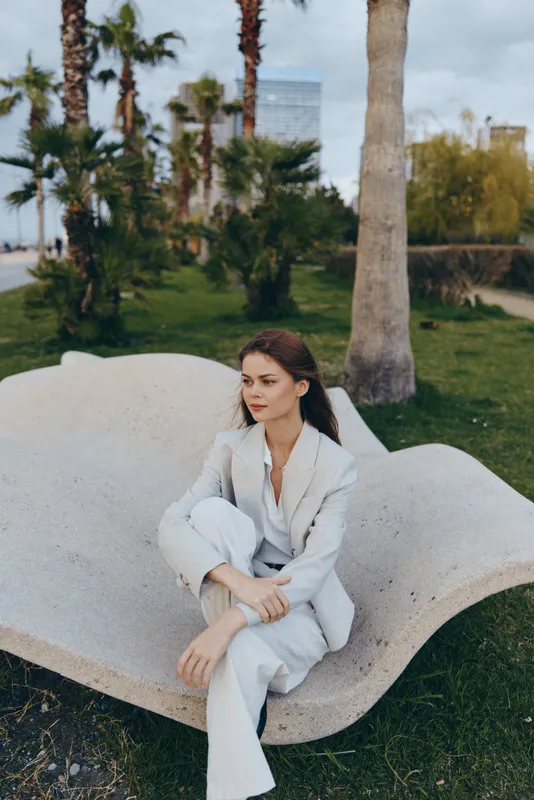 Business woman in a light suit sitting outdoors among palm trees, exuding confidence and professionalism in a scenic urban environment.
