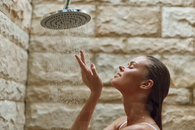 woman showering under rain showerhead with natural stone wall background, relaxed expression, wet hair, spa-like atmosphere, wellness concept