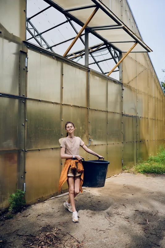 Woman walking outdoor beside a greenhouse carrying a bucket, smiling in casual outfit. Young adult female gardening task with sweater tied at waist and sneakers on dirt path.