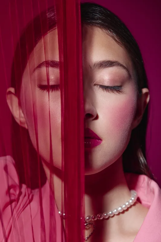 Portrait of young woman with closed eyes and bright makeup behind red sheer fabric wearing pearl necklace and pink blouse. Fashion beauty concept with elegant and artistic mood.