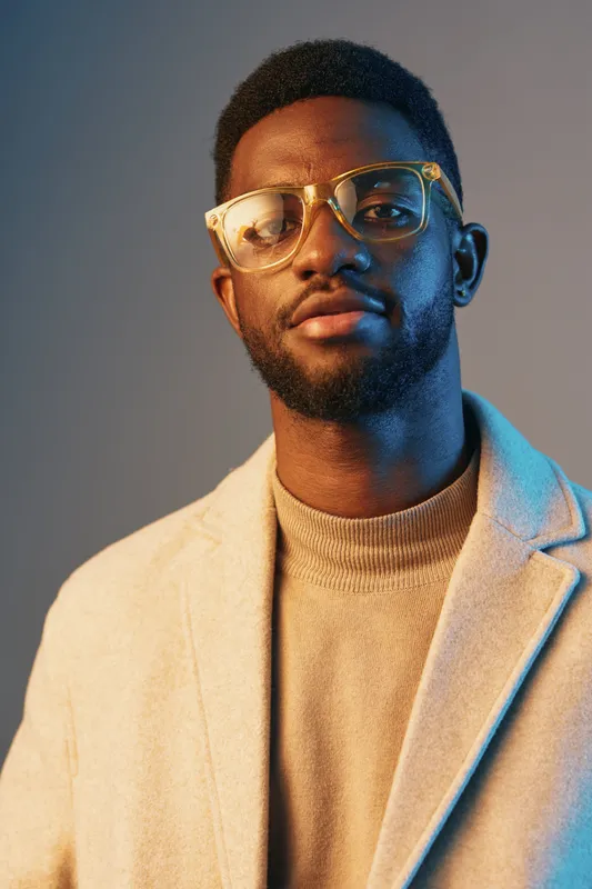 Stylish young Black man in glasses wearing a beige coat against a colorful background, showcasing confidence and modern fashion trends