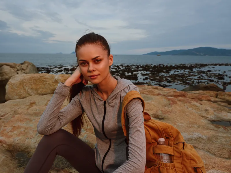 Young athletic woman in sportswear sitting on rocks near the sea with a contemplative expression, showcasing the beauty of active lifestyle and nature