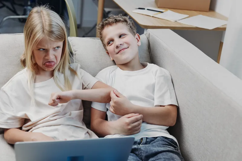 Annoyed girl and smiling boy sitting on couch, playful teasing and sibling rivalry captured in casual home setting with laptop nearby. Concept of family, childhood, and interaction.
