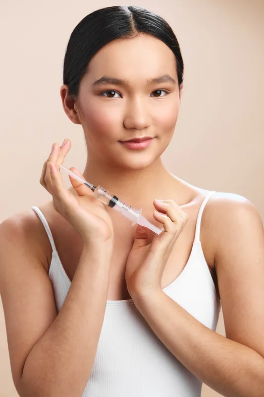 asian woman in a simple white top holds a syringe with calm confidence in a clinical setting, exuding professional care and reassuring presence for patients and caregivers, emphasizing safety,