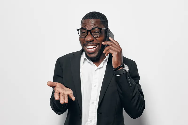 African American man with dark skin wearing glasses and black suit talking on phone with expressive gesture isolated on white background in studio.