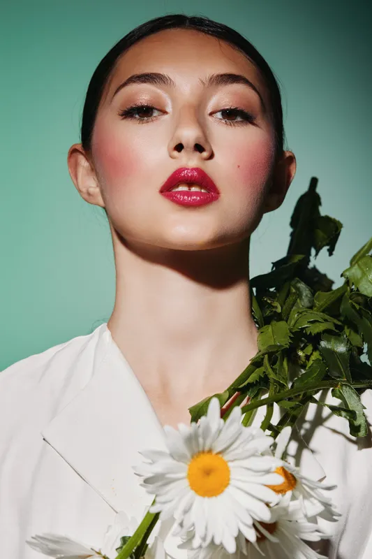 Close-up of a young woman with glossy red lips and perfect skin holding a white daisy with greenery on a pastel green background. Fashion beauty creative studio portrait.