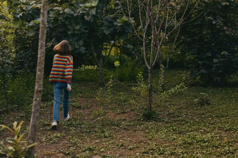Child walking in forest in striped sweater and jeans, back view of kid exploring nature among trees and shrubs, outdoor portrait of youthful curiosity in a quiet green landscape