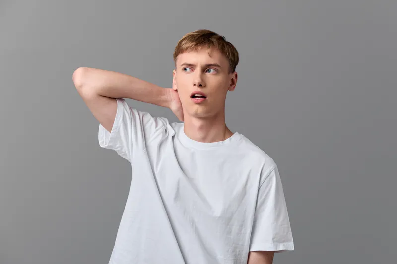 Young man in a white tee scratches his neck with a puzzled expression, standing in a neutral gray studio, casual pose and uncertain mood captured in a simple shot