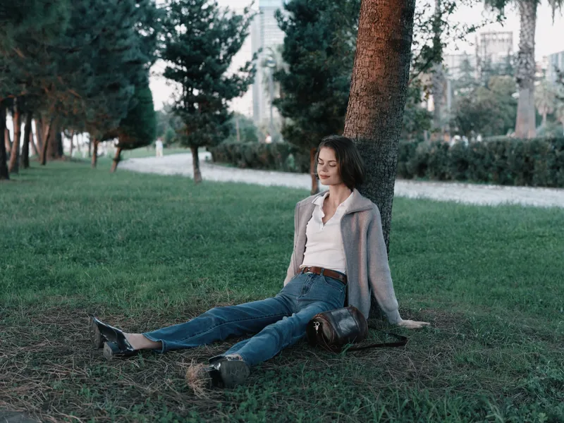A thoughtful woman sits beneath a tree in a quiet park, dressed in casual attire. She rests on the grass, gazing ahead, conveying calmness and reflection in a natural outdoor setting.