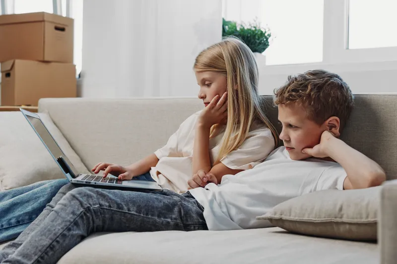 Two children sit on a beige sofa using a laptop together in a bright living room. The girl and boy focus on the screen with casual clothes and relaxed posture, concept for learning and technology.