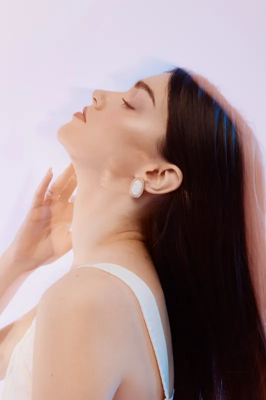 Young woman beauty fashion model with long dark hair poses with closed eyes and elegant earrings on soft pastel background. Concept of style and skincare.