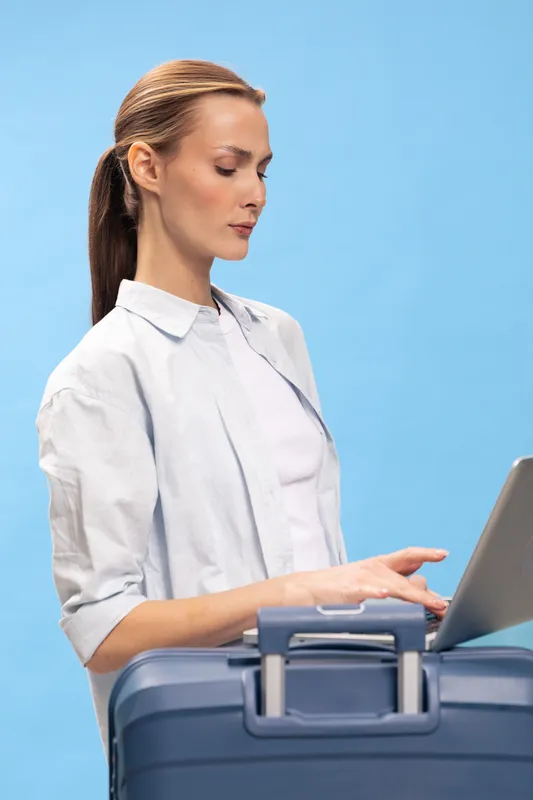 Focused woman with long hair types on laptop while standing behind suitcase against blue background. Concept of travel planning, business trip, and modern digital lifestyle.