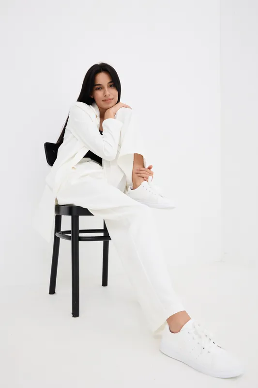 Young woman sitting on black chair wearing white suit and sneakers, posing in minimalistic bright studio, casual style and relaxed confident mood.