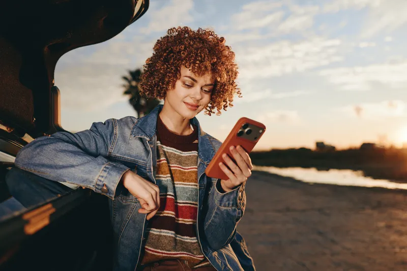 Woman lifestyle sunset smartphone car road smiling curly hair, jeans denim jacket striped shirt, holding phone near parked vehicle, warm evening sunlight, relaxed travel outdoor candid portrait