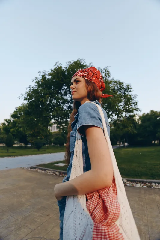 lifestyle woman in modern boho-western denim street style with a red bandana and tote bag, captured in warm film color tones outdoors in a park setting