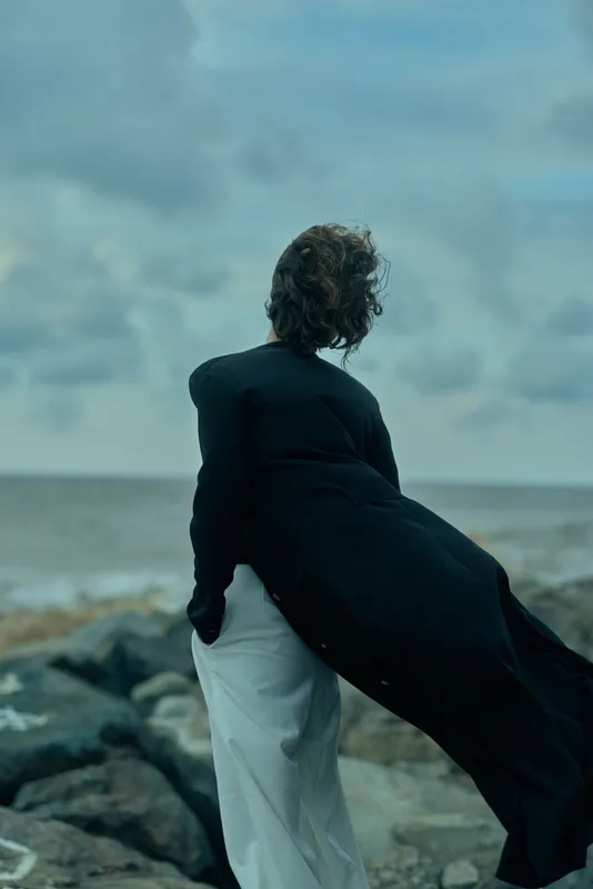 Woman standing on a rocky beach, long black coat billowing in the wind, facing the sea under a cloudy sky, creating a moody fashion moment