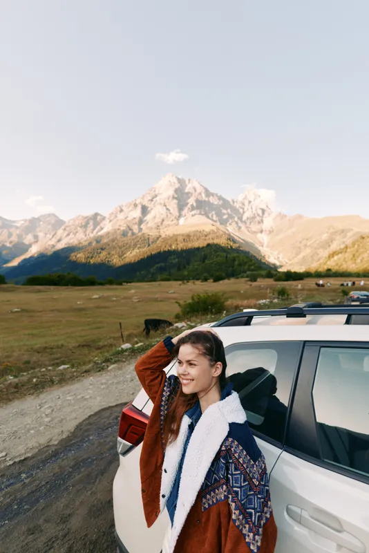 Woman car mountains travel portrait roadtrip landscape nature young woman leaning on white SUV smiling with jacket, scenic countryside road and panoramic mountain view under open sky.