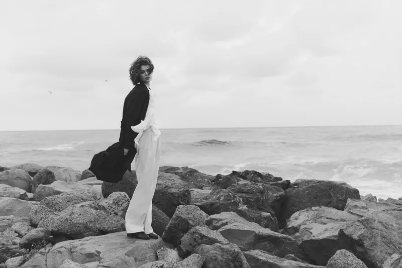 Woman standing on rocky shore by the sea, wind in her hair, flowing dress, monochrome mood, dramatic coastline outdoors with atmosphere and movement