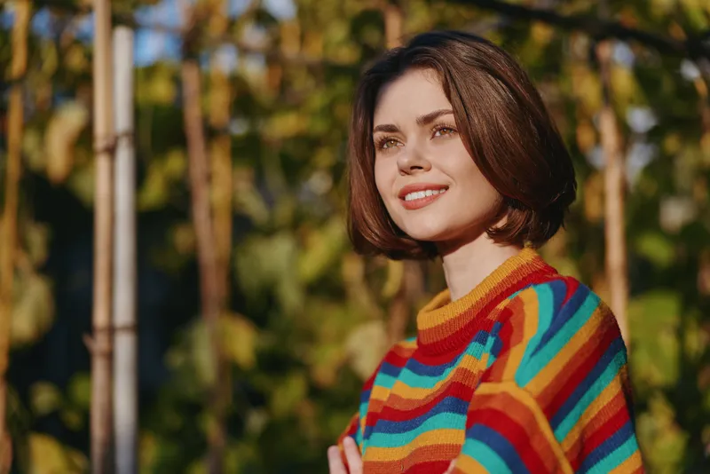 Woman portrait smile in colorful sweater outdoors in warm sunlight, short brown bob haircut, shallow depth of field, natural green bokeh background and relaxed confident expression.