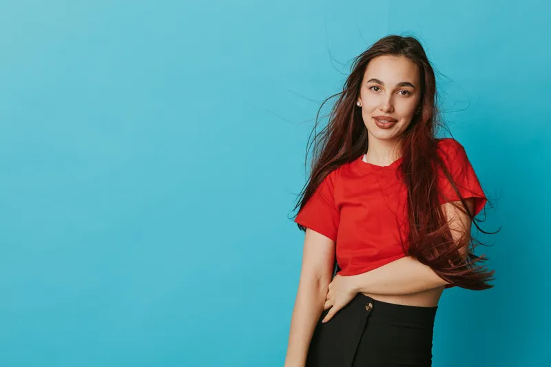 Young woman with long hair wearing red t shirt and black pants posing against blue background smiling and looking at camera in casual style with natural makeup and relaxed posture.