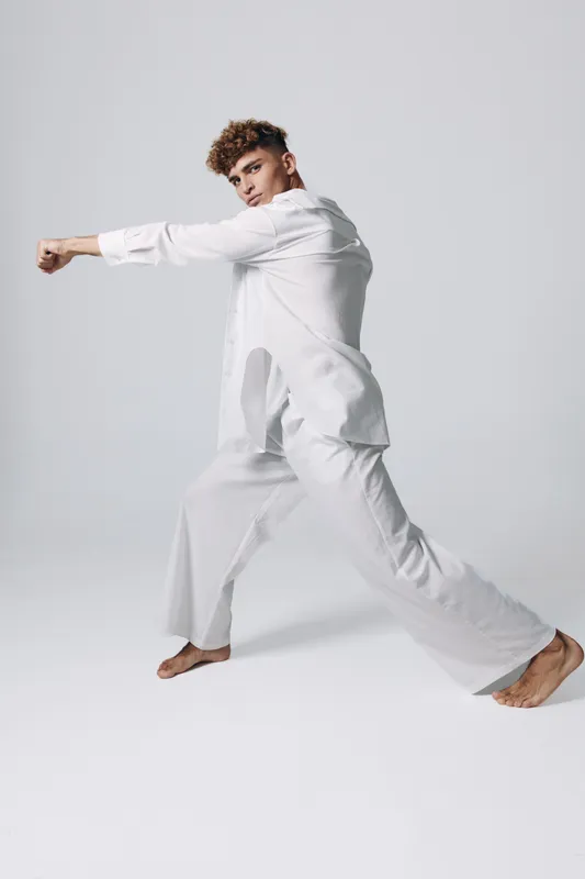Young man in white clothes practicing martial arts with focused expression and extended punch in studio with plain background and barefoot stance.