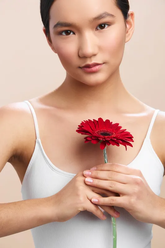 asian woman holds a vivid red flower close to her chest in a simple studio setting. she wears a white tank top, soft lighting, calm expression, and a serene, graceful mood.