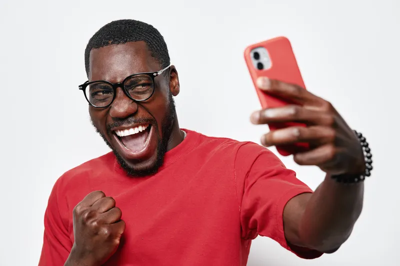 African American man with dark skin and glasses taking selfie with smartphone, smiling and celebrating success in casual red shirt on white background.