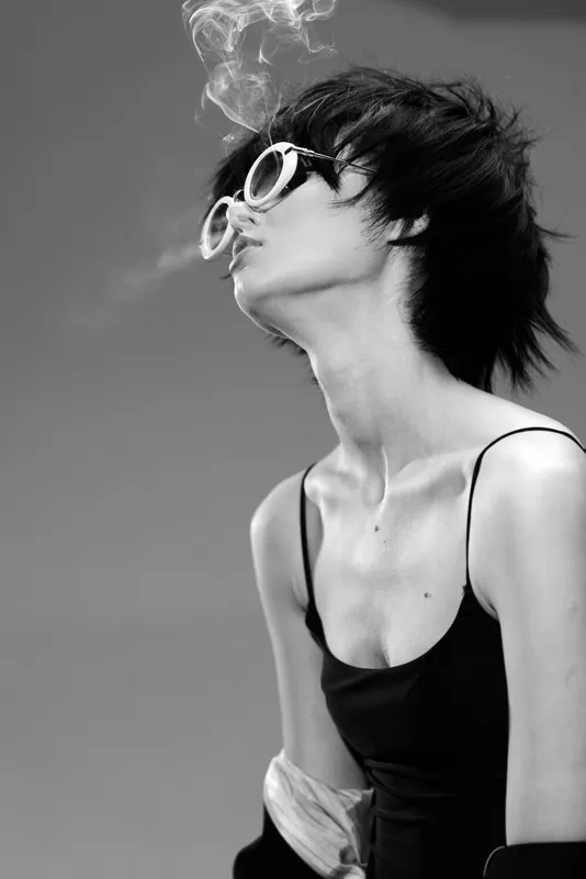 Black and white portrait of a young woman smoking, wearing sunglasses and a tank top, showing a rebellious and stylish attitude.