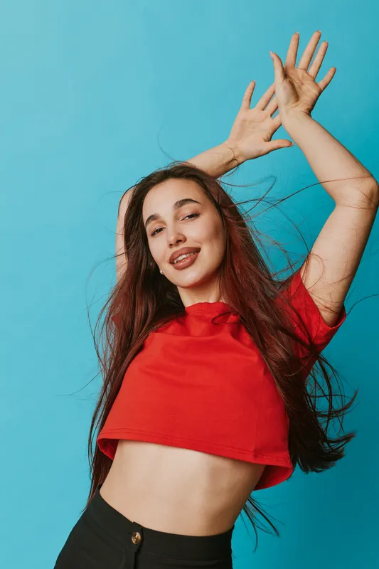 Young woman in red crop top dancing with hands raised smiling long hair flowing against blue background in studio. Casual style, joyful expression and motion captured in portrait.