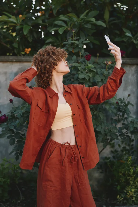Cheerful woman outdoors, wearing rust outfit, posing for a selfie, warm sunlight on her smile, relaxed posture among green plants, natural tones, candid lifestyle moment in frame