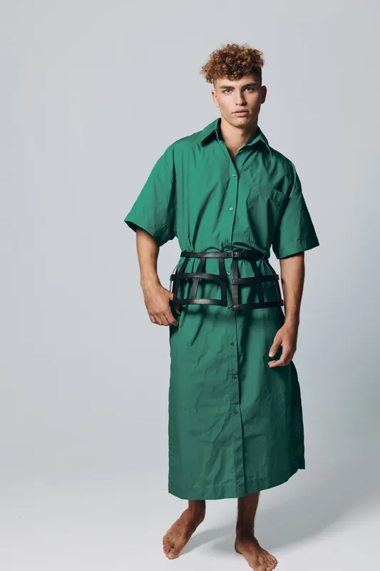 Young man with curly hair wearing a green oversized shirt dress and a black harness belt standing barefoot on a plain background in studio lighting.