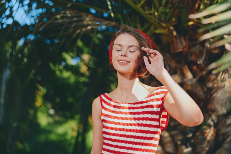 A cheerful young woman stands outdoors in bright sunlight, smiling with closed eyes, wearing a red and white striped tank top, enjoying a warm, sunny day amid lush greenery.