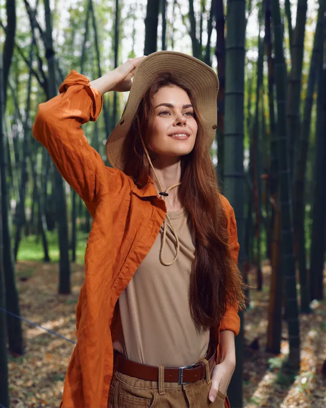 Woman in hat and longhair smiling in bamboo forest, standing with hand on brim, casual orange jacket and neutral blouse, nature walk, serene outdoor portrait.