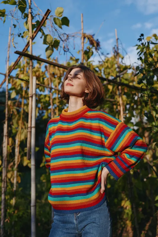 Young woman in her mid 20s wearing a colorful striped sweater and casual outfit, smiling and standing in an outdoor garden among trellis vines under warm sunlight.
