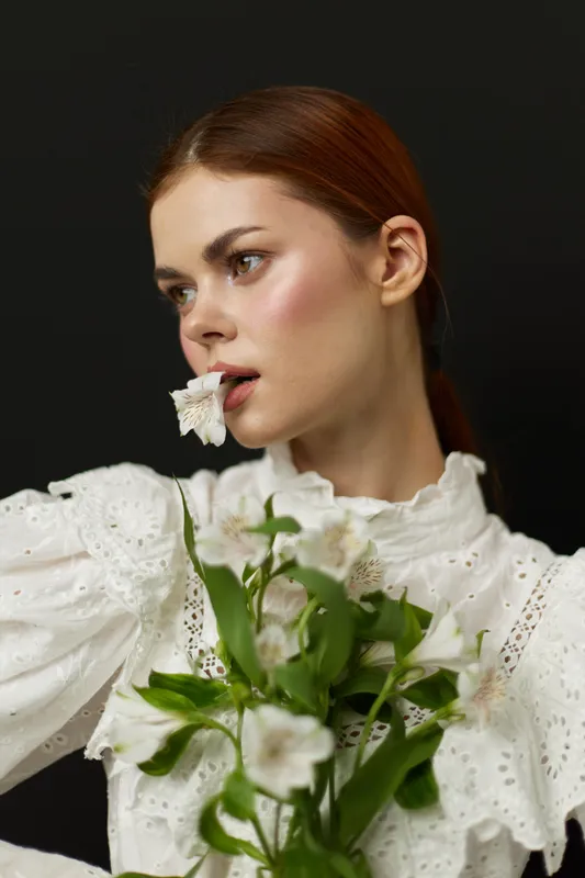 Elegant woman in white lace blouse holding flowers, exuding serenity and beauty against a dark background, perfect for fashion and beauty concepts