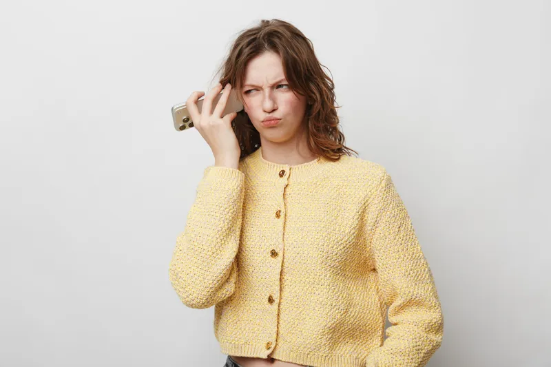 Confused young woman in yellow sweater holds smartphone to her ear, frowning and looking puzzled. Concept of misunderstanding, communication problems, and frustration with technology.