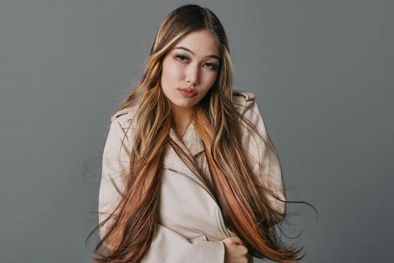 Young woman with long hair wearing light beige coat standing with folded arms looking at camera on gray background. Studio portrait with serious facial expression and casual style.