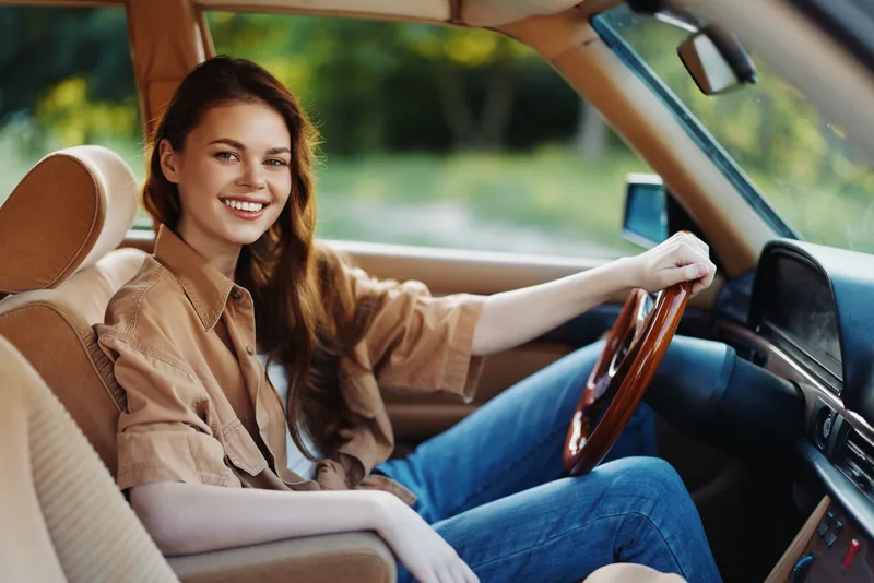 Happy young woman driving a vintage car, wearing a casual outfit, showcasing a joyful expression in a natural green background.