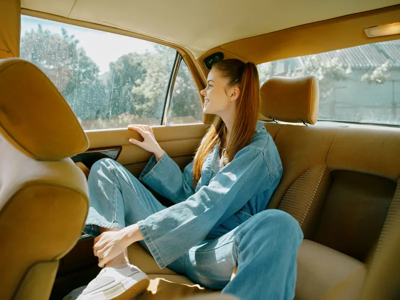 Joyful young woman enjoying a sunny ride in a vintage car, dressed in casual denim, showcasing happiness and relaxation amid warm colors and a bright backdrop.