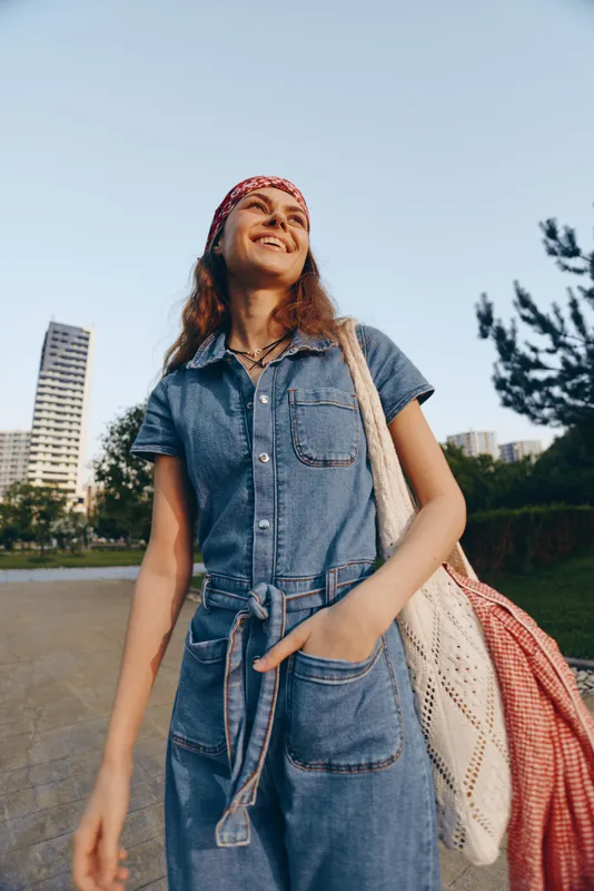 smiling woman in denim jumpsuit enjoying sunny day outdoors with casual red headband and tote bag in urban park setting