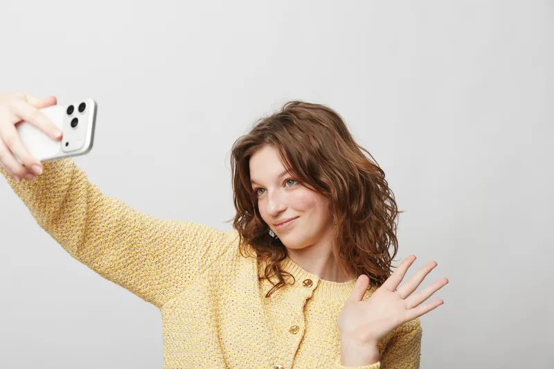 Young woman with curly hair wearing yellow sweater takes selfie with smartphone and waves hand. Bright studio background. Concept of social media, communication, and casual lifestyle.