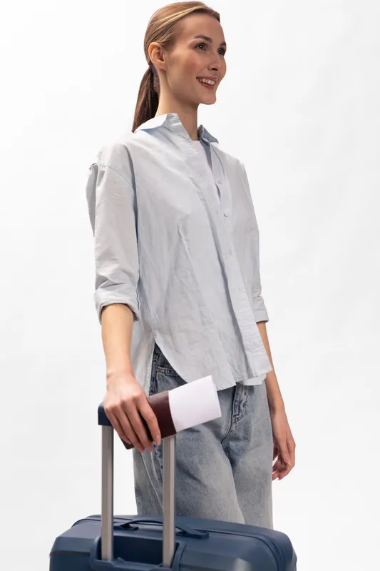 Young woman traveler stands with suitcase and passport in hand, smiling and looking ahead. Casual outfit and bright background create a positive travel concept and adventure mood.