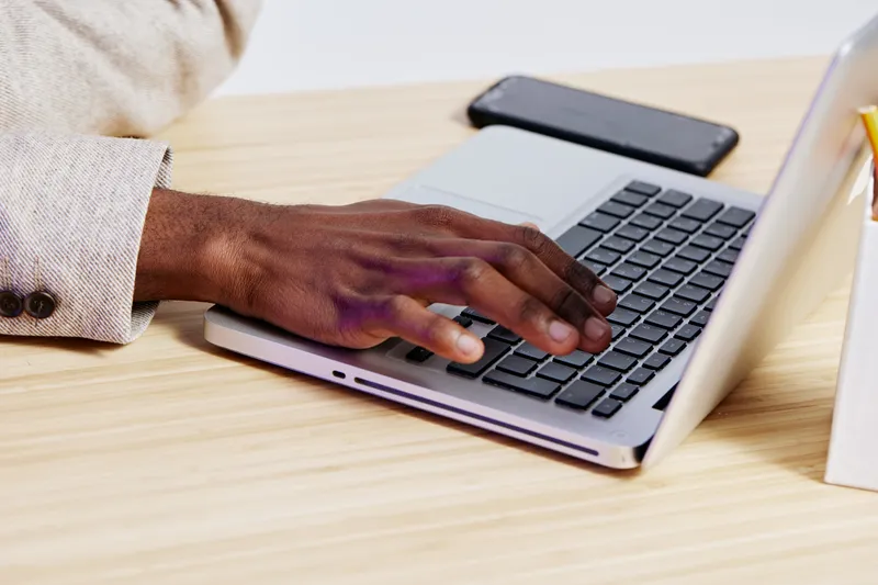 Close-up of a person's hand typing on a laptop keyboard at a wooden desk, with a smartphone and notepad nearby, ideal for workplace or remote work themes, modern office environment, technology use, pr