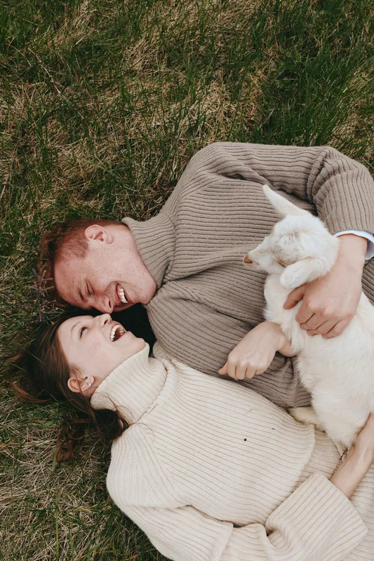 Smiling couple lying on grass with a curious goat kid, embracing and laughing in warm natural light. Cozy sweaters, outdoor vibe, intimate lifestyle moment with playful animal companionship.