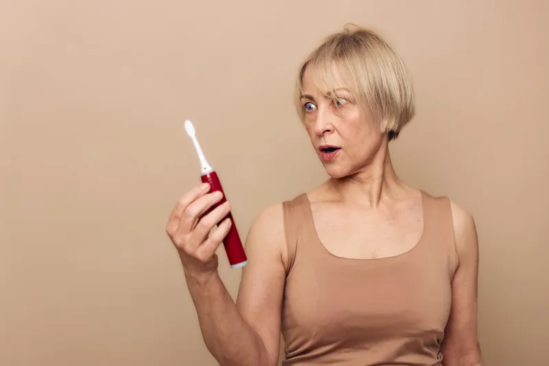 Surprised mature woman in beige top looks at electric toothbrush with wide eyes. Neutral background emphasizes expression of curiosity and disbelief. Concept of daily hygiene and unexpected moments.