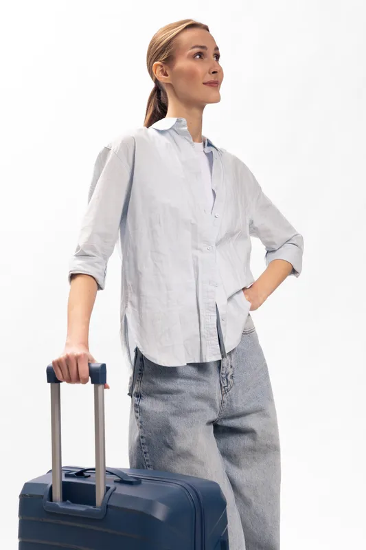 Young woman traveler with blue suitcase stands confident in casual white shirt and loose pants. Concept of travel, tourism, vacation, and independence on white background.