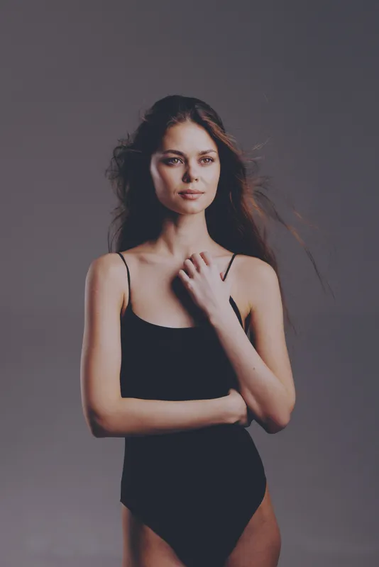 Young woman in black bodysuit posing confidently with natural makeup and loose hair on gray background